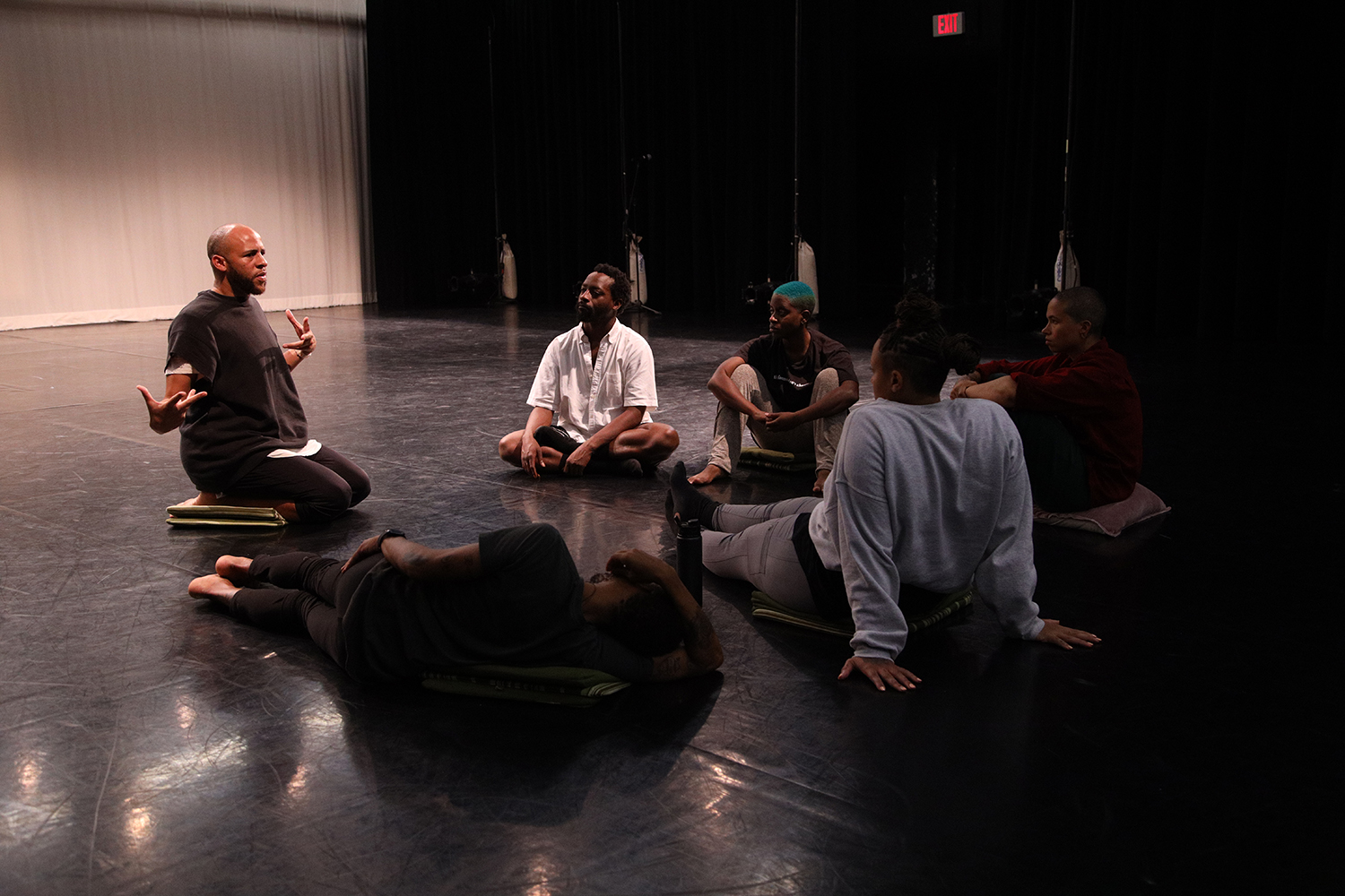 Five people sit or lay on cushions in a circle on a black floor, engaged in a rehearsal discussion, led by a person sitting upright and gesturing surrounded by black curtains and a white scrim.