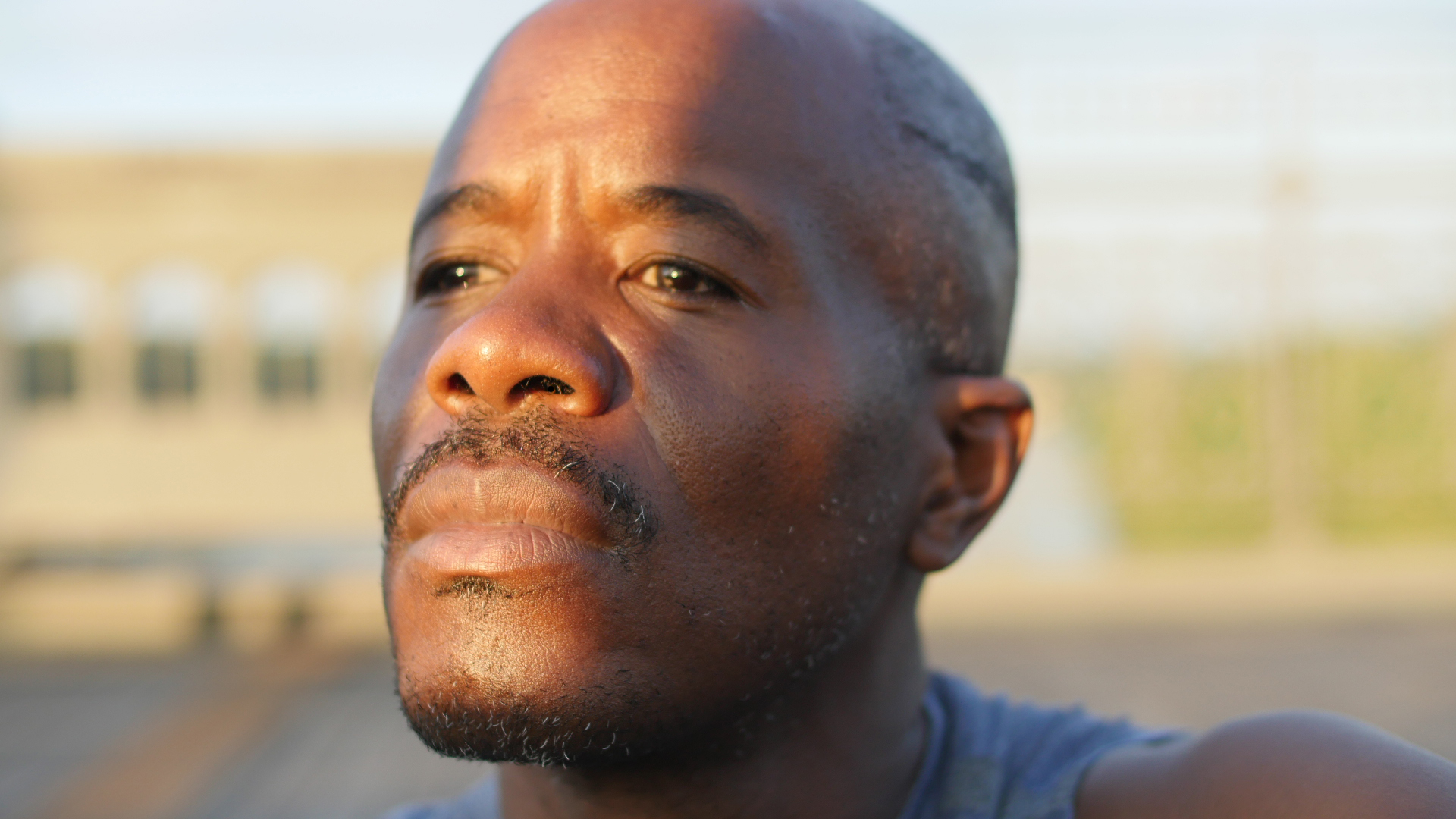 A man stares forward, seemingly deep in thought. His face is very close to the camera. Behind him, a natural area is out of focus. 
