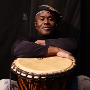 A man wearing a hat poses with his arms crossed over an African Drum