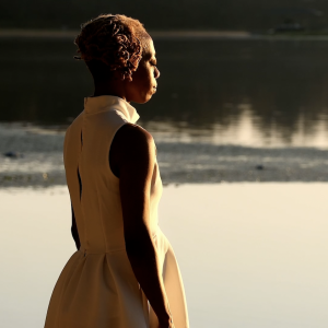 Kehinde Ishangi is wearing a light yellow dress and looks mournfully out at a lake.