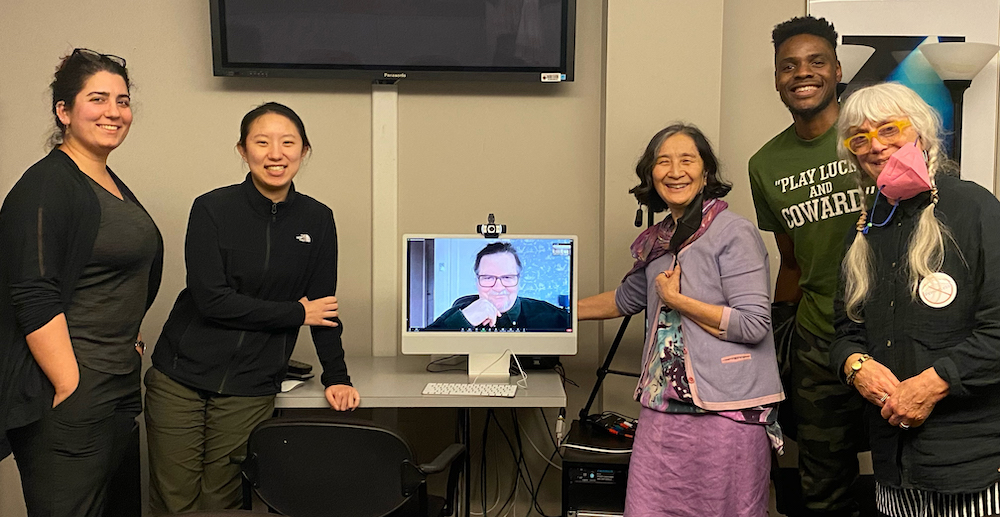 A group poses for a photo around a computer screen, where a man is on a video call. (Stephanie Battle)