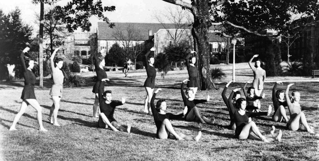 A black and white photo of young women performing in a grassy area on a college campus. 