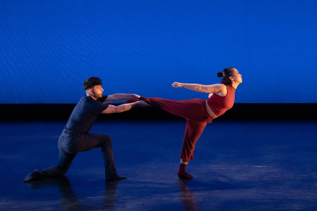 Two dancers, a man and a woman, perform in front of a blue backdrop. 