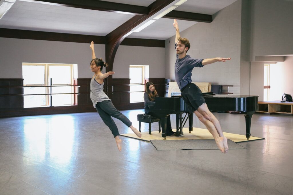 Dancers are leaping in a studio while someone plays the piano in the background
