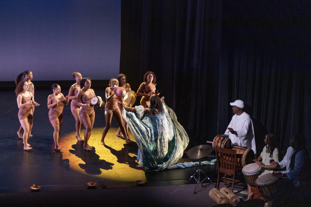 Dancers in colorful costumes hold percussion instruments. A man in a white outfit is playing an Africal drum.
