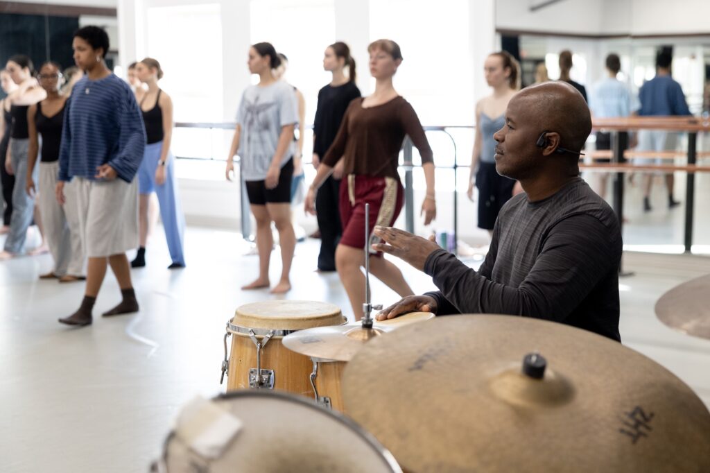 A man sits at a drum set with dancers in the background