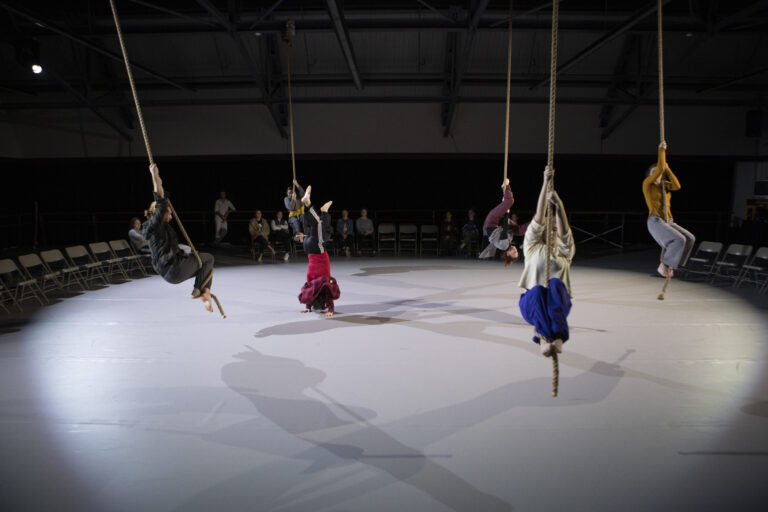 five people in a circus type dancing hanging from rope on circus stage with chairs around in circle