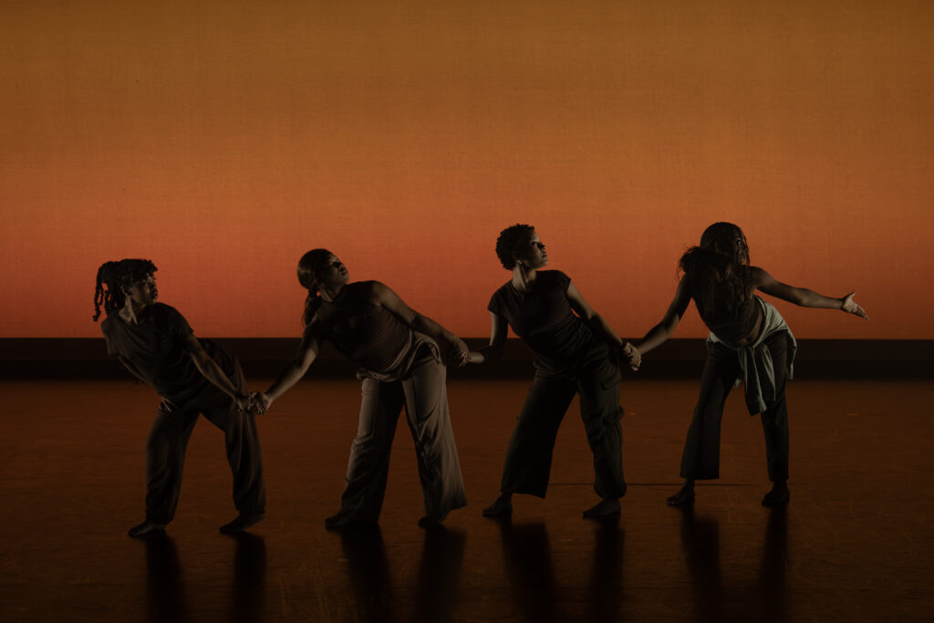 Four dancers silhouetted in a line with joined hands- reaching to the right