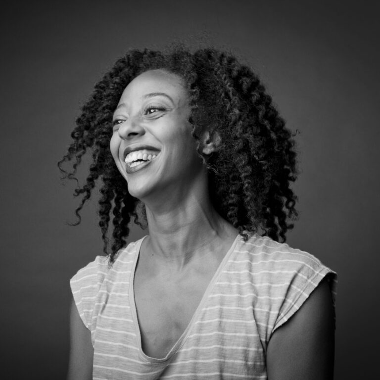ID: A black-and-white image of queer Trinidadian femme, Fana Fraser, laughing. Her upper body is angled away from the camera lens as she looks into the distance with glee. She is comfortably dressed in a striped cap sleeved v-neck t-shirt. Their eyes shine with a smile that is broad, open mouthed and baring teeth. Her hair falls at shoulder length, unraveled in a natural two strand twist out. The photo is a portrait by Whitney Browne from a 2014 studio session in New York City.