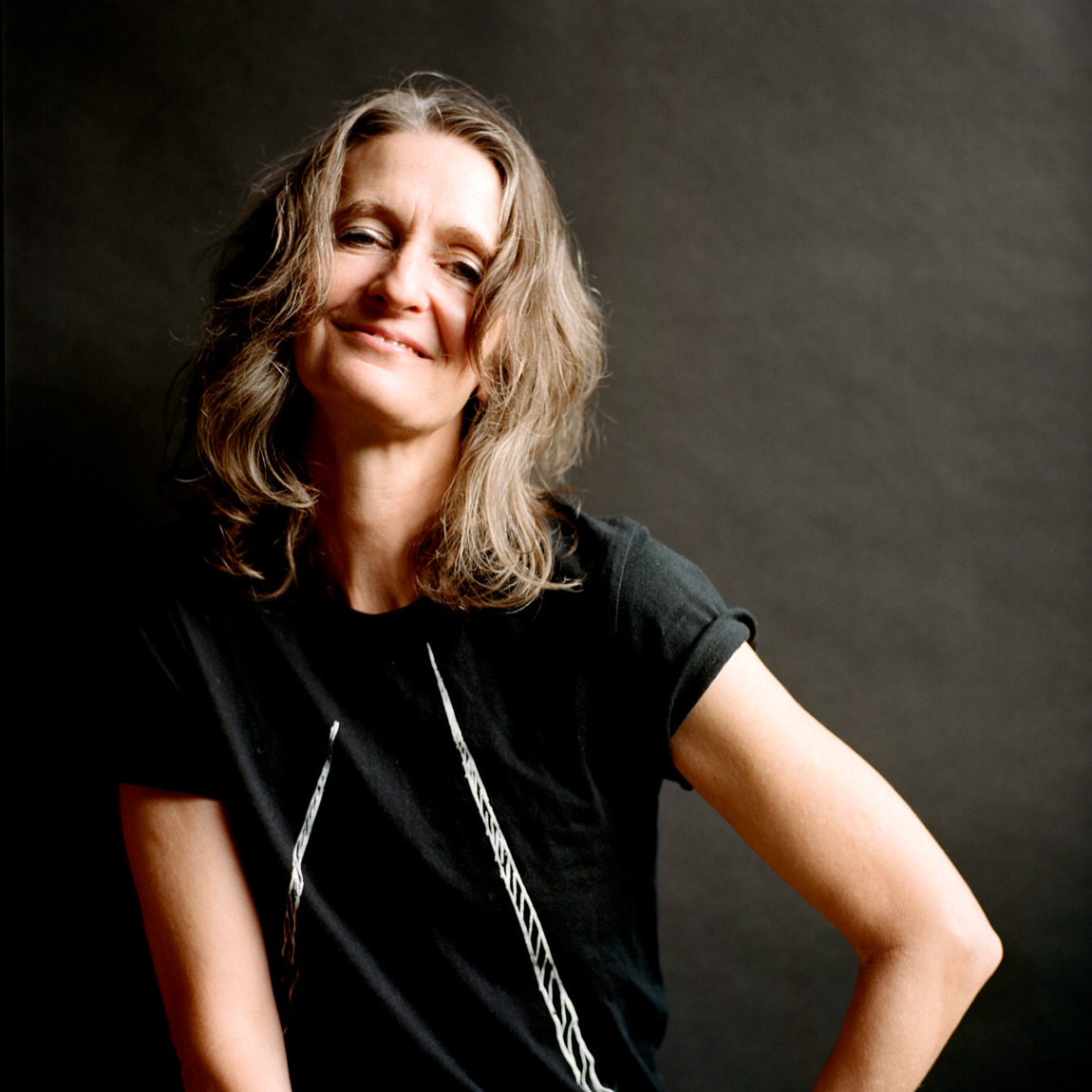 A woman with long, wavy brown hair smiles gently at the camera. She is wearing a black t-shirt with a white graphic design.