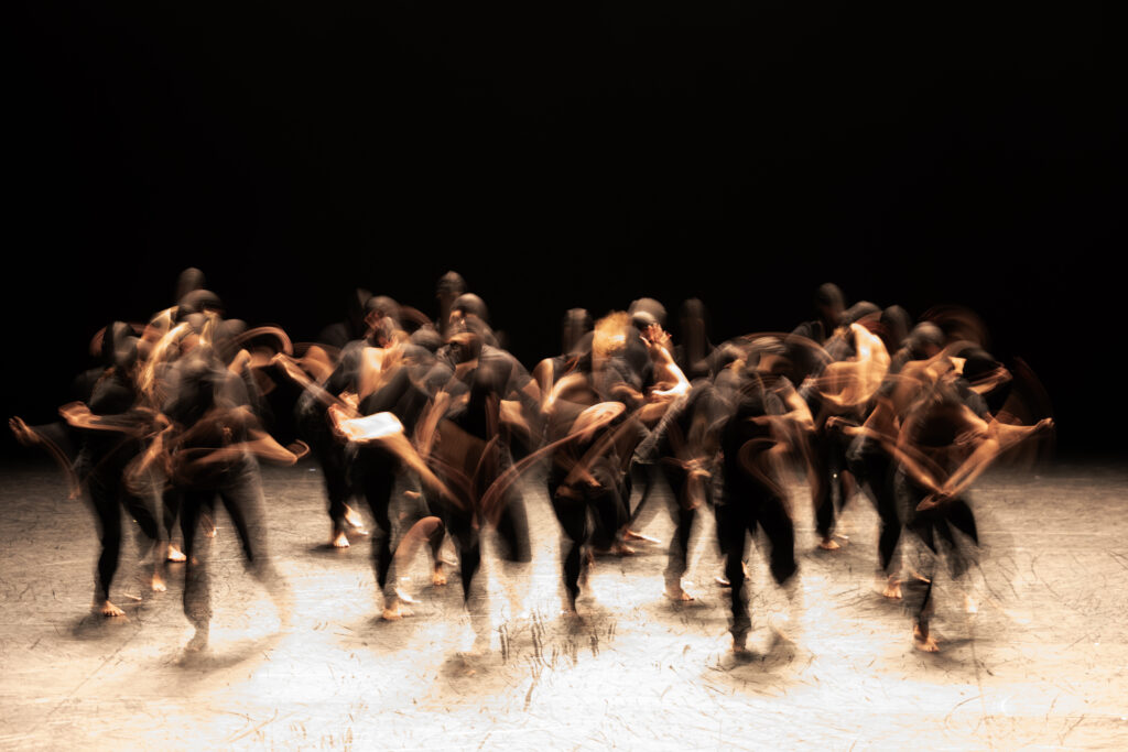 A long exposure photo of a large ensemble of dancers, showing blurred lines of light and color