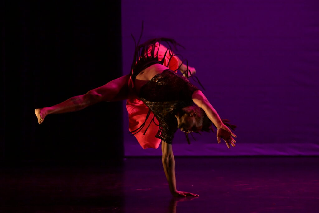 A dancer wearing red upside down supported by only one hand