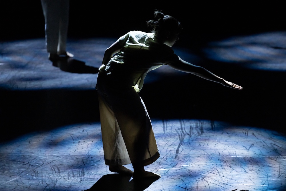 A dancer standing in a pool of light reaching out to the right
