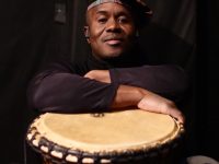 A man wearing a hat poses with his arms crossed over an African Drum